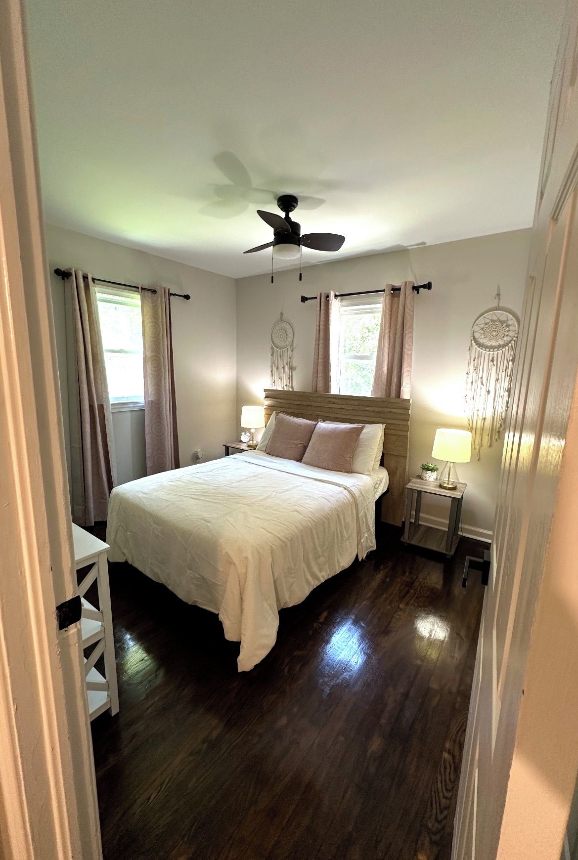 A bedroom with a bed, a ceiling fan, a nightstand with a lamp, and a dreamcatcher on the wall.