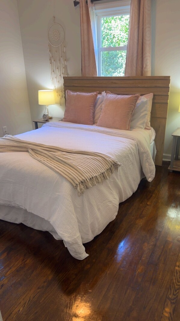 Una cama bien hecha con una manta y almohadas color beige, una lámpara de noche y un atrapasueños en la pared.