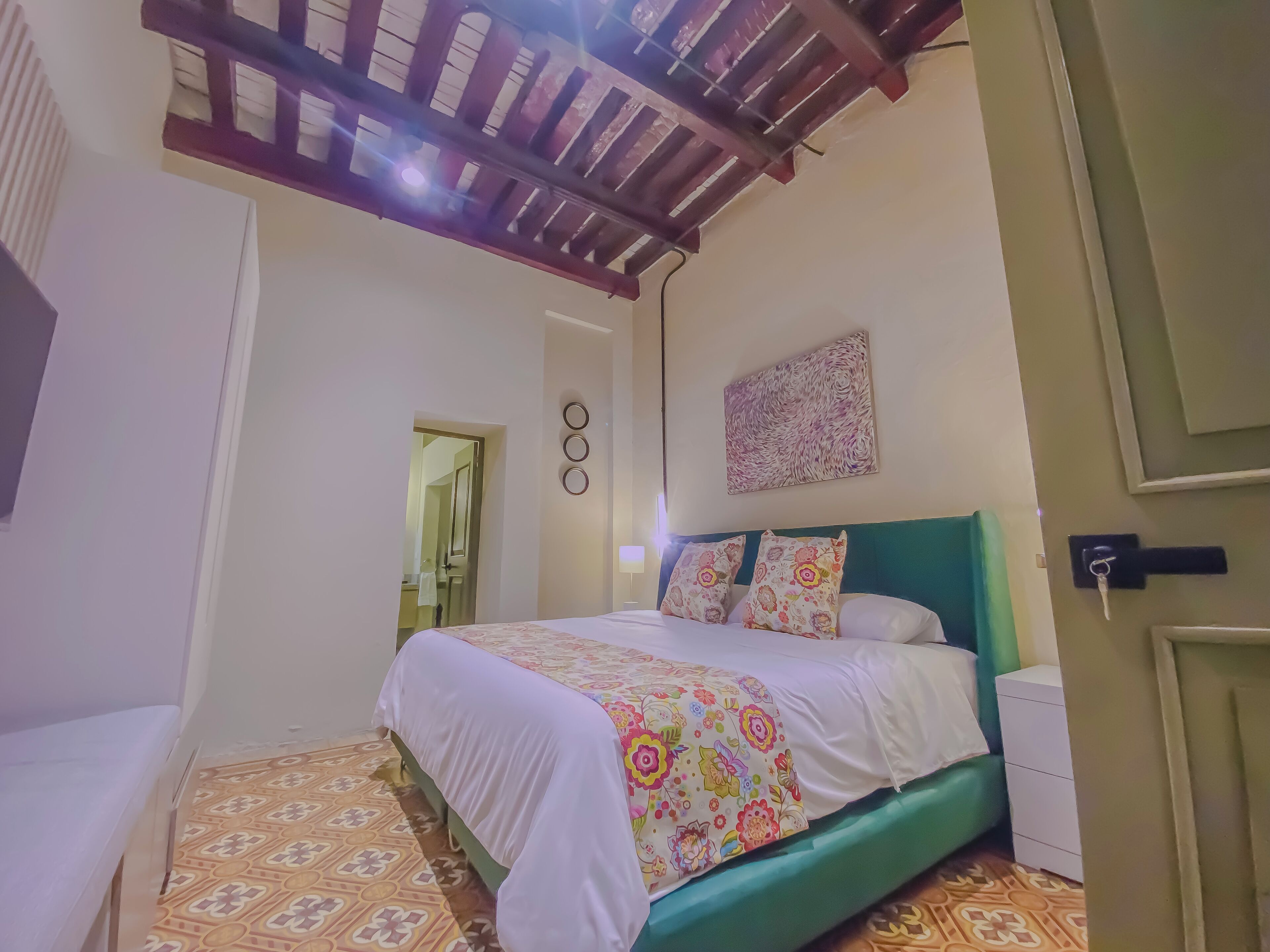 A hotel room with a bed, a green headboard, a patterned floor, a wall-mounted artwork, and a door with a key in the lock.