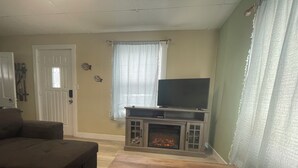 A living room with a fireplace, a television on a wooden stand, a sofa, and a window with curtains. - The Purple Door -FRESHLY UPGRADED AIR CONDITIONING NOW AVAILABLE- (McGill)