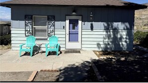 A small, single-story house with a dark roof and light siding. There are two turquoise chairs outside, a small table, and a door with a window. - The Purple Door -FRESHLY UPGRADED AIR CONDITIONING NOW AVAILABLE- (McGill)