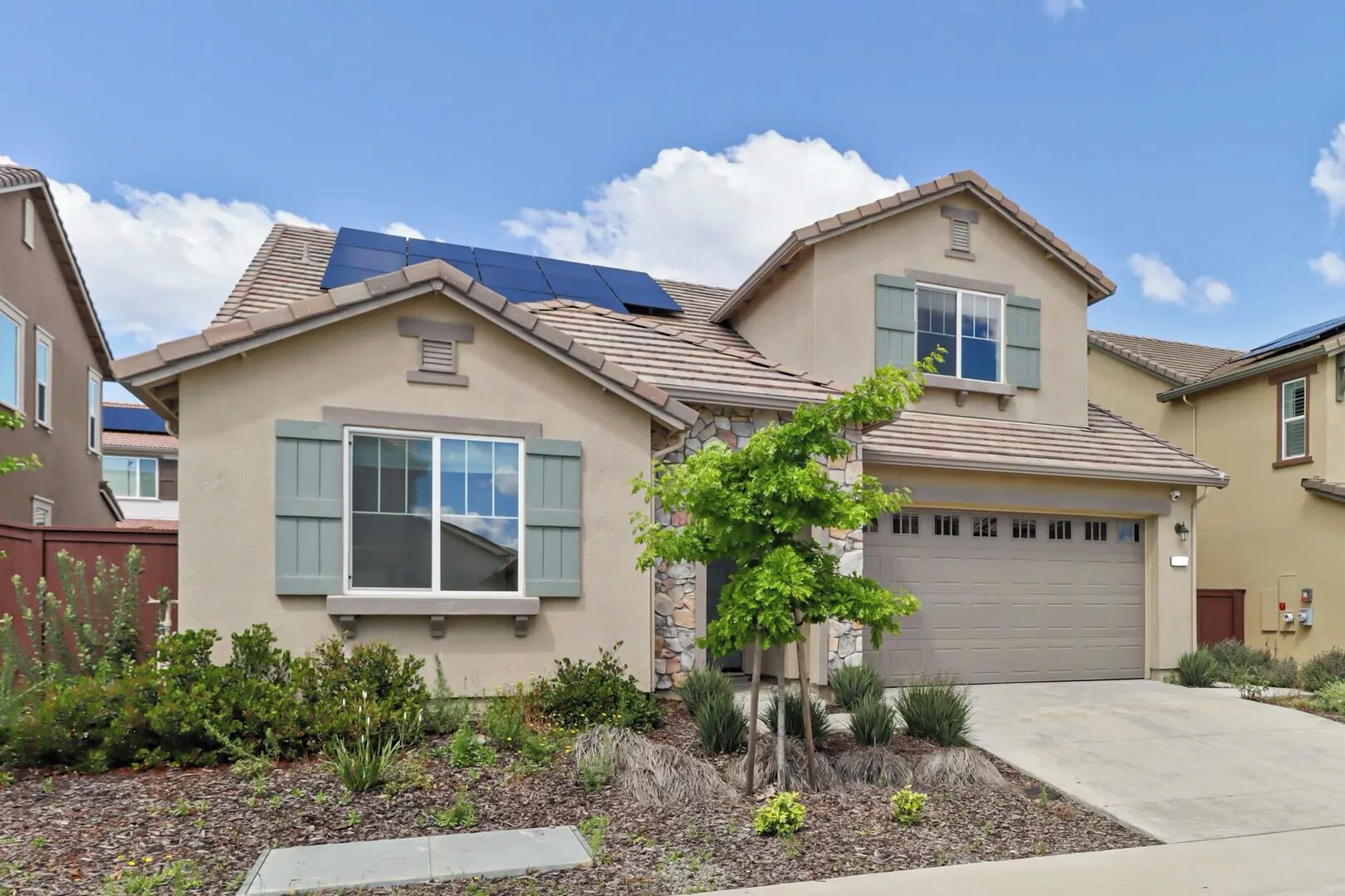 A residential house with a garage, a small tree in the front yard, and solar panels on the roof.