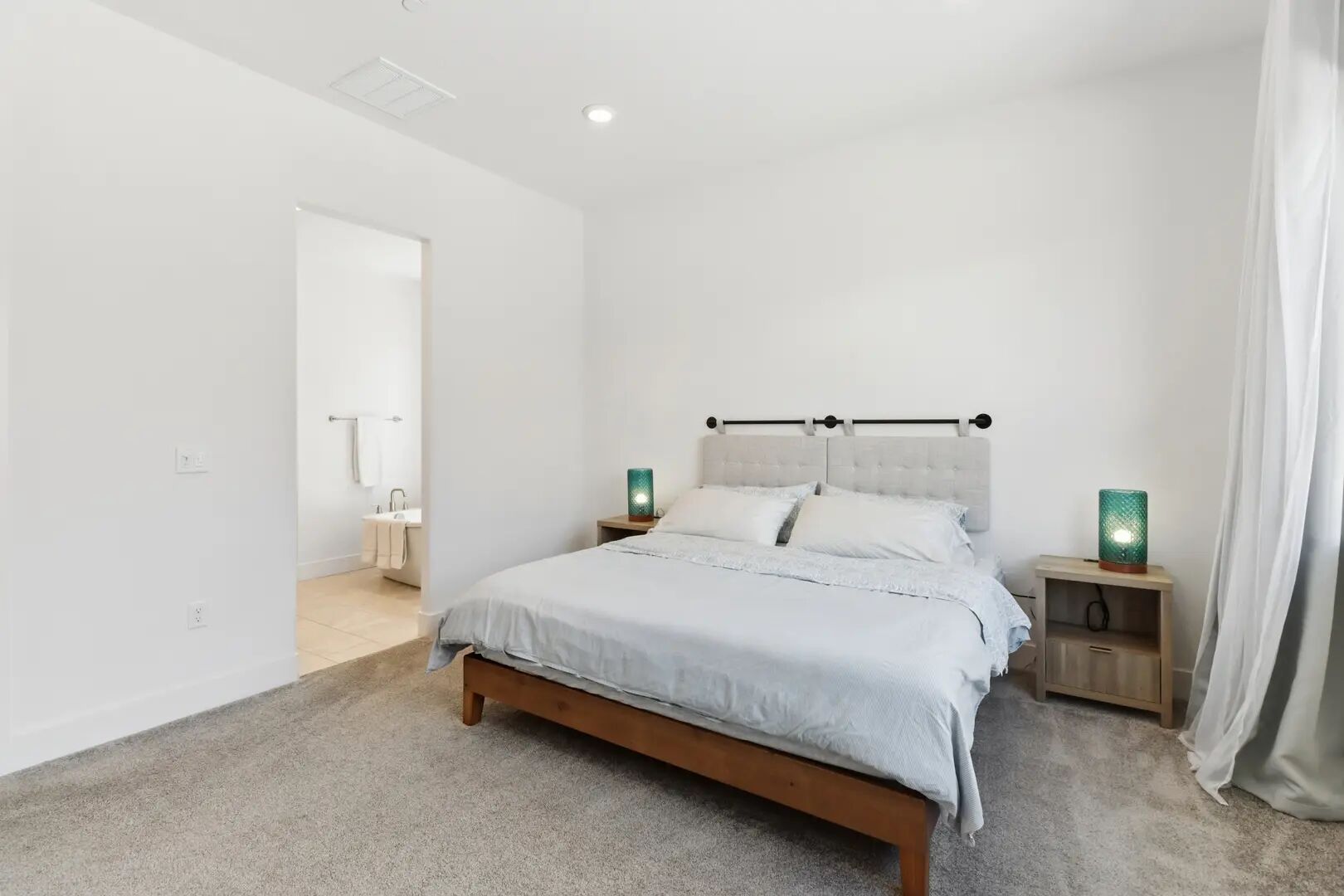 A bedroom with a bed, bedside tables, a nightstand, a bathroom with a sink and mirror, and a door leading to another room.