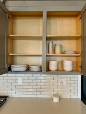 Una cocina con salpicadero de azulejos blancos, una encimera con un dispositivo blanco y un armario con estantes que contienen platos y tazones.