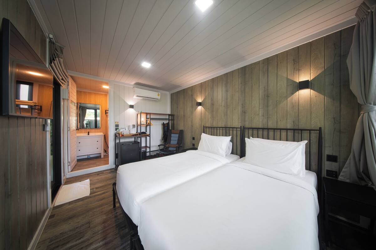 A hotel room with a wooden floor, a bed with white linens, a desk with a chair, a mirror, and a window with curtains.