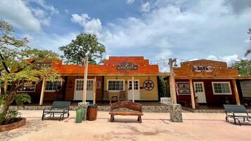Un edificio con temática del Viejo Oeste, que incluye una oficina de alguacil y un banco, con mesas al aire libre y un árbol.