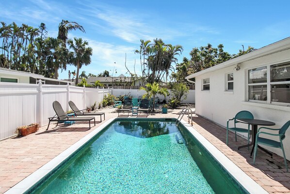 A rectangular swimming pool with a white deck, surrounded by lounge chairs and a small table.