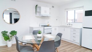 A modern kitchen with white cabinets, a gas stove, and a window. There is a round mirror on the wall and two potted plants on the floor. - Spacious & Cosy Self-Contained 2 Bedroom Apartment (Rivervale)