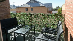 A patio with a glass table and two chairs, a brick wall, and a fence with climbing plants. - Spacious & Cosy Self-Contained 2 Bedroom Apartment (Rivervale)