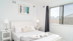 A white bedroom with a bed, two bedside tables, a window with curtains, and a framed picture on the wall. - Spacious & Cosy Self-Contained 2 Bedroom Apartment (Rivervale)