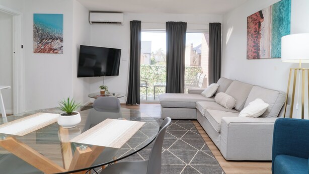 A modern living room with a glass dining table, grey sofa, and a flat-screen TV mounted on the wall. - Spacious & Cosy Self-Contained 2 Bedroom Apartment (Rivervale)