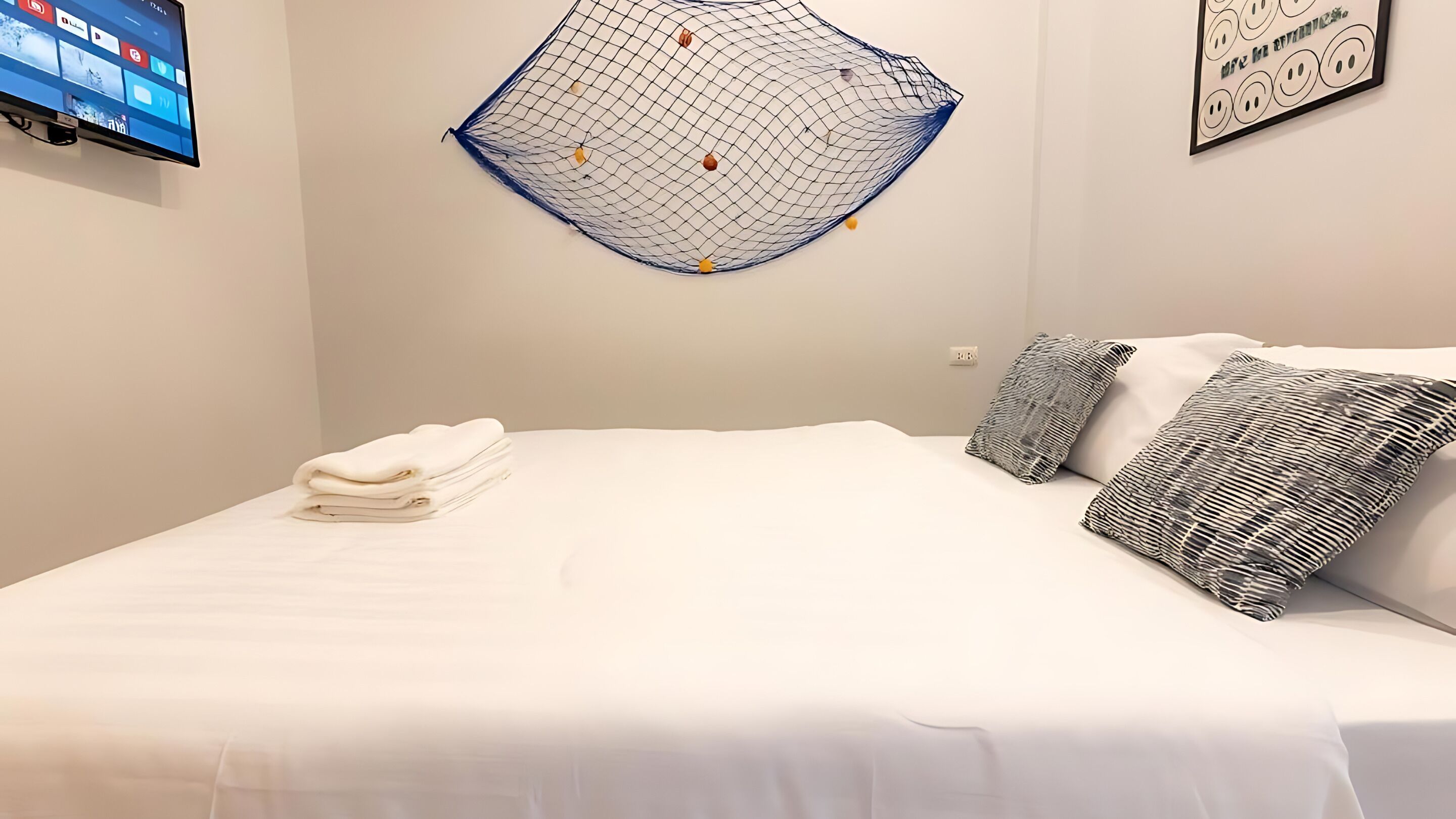 Un lit bien fait, avec une literie blanche et deux coussins à motifs, un téléviseur fixé au mur et une décoration murale.