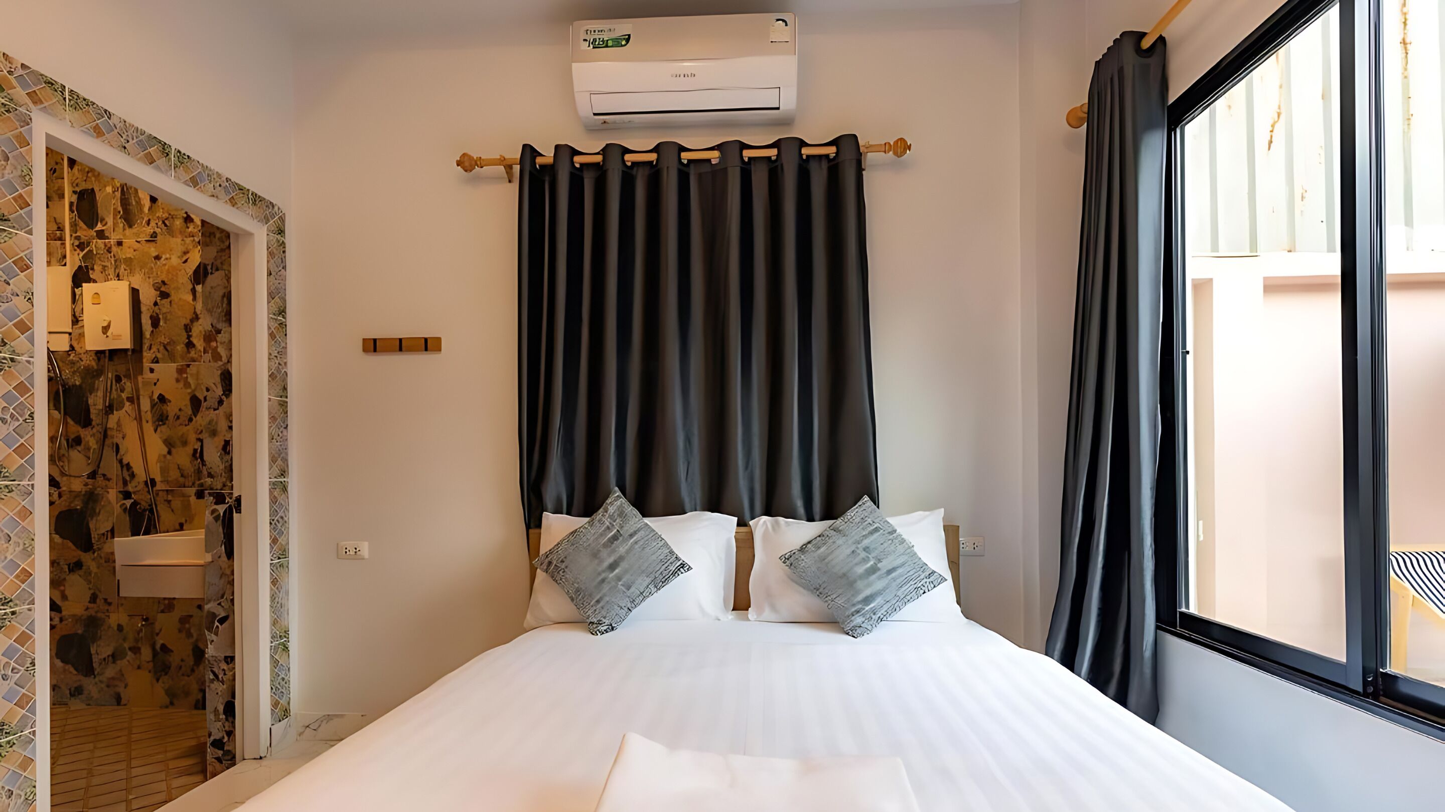 Ein Schlafzimmer mit einem Bett, zwei Kissen, einem Fenster mit Vorhängen, einer Klimaanlage und einem Badezimmer mit Dusche.