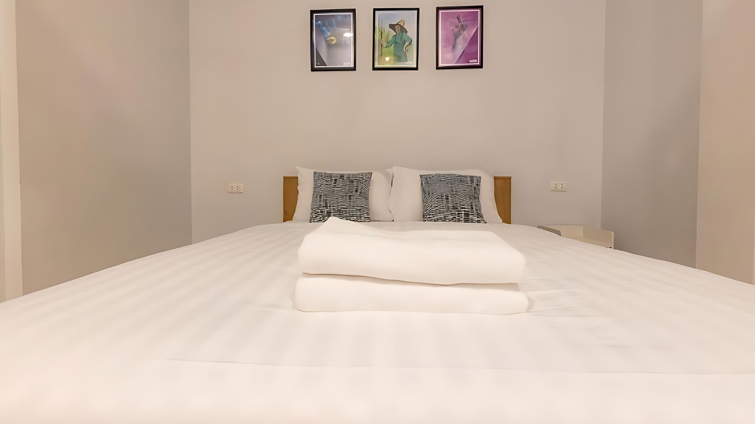 Un lit bien fait, avec du linge de lit blanc et deux coussins à motifs, une tête de lit en bois et trois œuvres d’art encadrées accrochées au mur au-dessus.