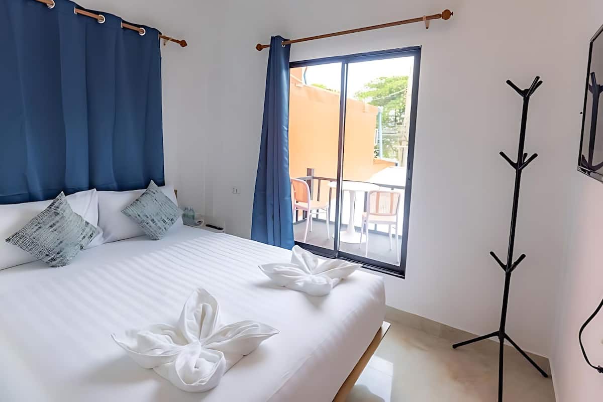 A hotel room with a bed, blue curtains, a balcony with chairs, and a coat rack.