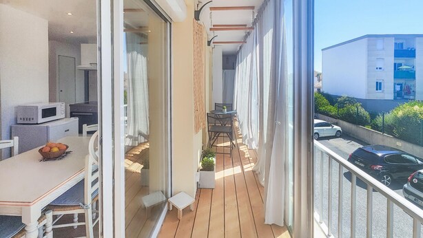 A balcony with a wooden floor, a dining table with chairs, a small table with a plant, and a view of a parking area and buildings. - 1 bedroom nice apartment in Palavas-Les-Flots (Palavas-Les-Flots)