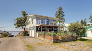 A two-story house with a balcony, a fenced garden, and a clear view of the water in the background. - Sunset + Ocean Views: Serene Camano Escape (Camano)