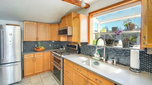 A modern kitchen with wooden cabinets, a stainless steel refrigerator, a built-in oven, and a large window with potted plants. - Sunset + Ocean Views: Serene Camano Escape (Camano)