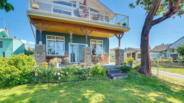 A two-story house with a balcony, stone foundation, and a well-maintained lawn. - Sunset + Ocean Views: Serene Camano Escape (Camano)