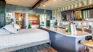 A modern bedroom with a large bed, a small island desk, and a kitchenette. - Fallbrook Airplane Home w/ Fire Pit & Mtn Views! (Fallbrook)