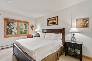 A bedroom with a large bed, two bedside tables with lamps, a window with a view of trees, and two wall-mounted paintings.
