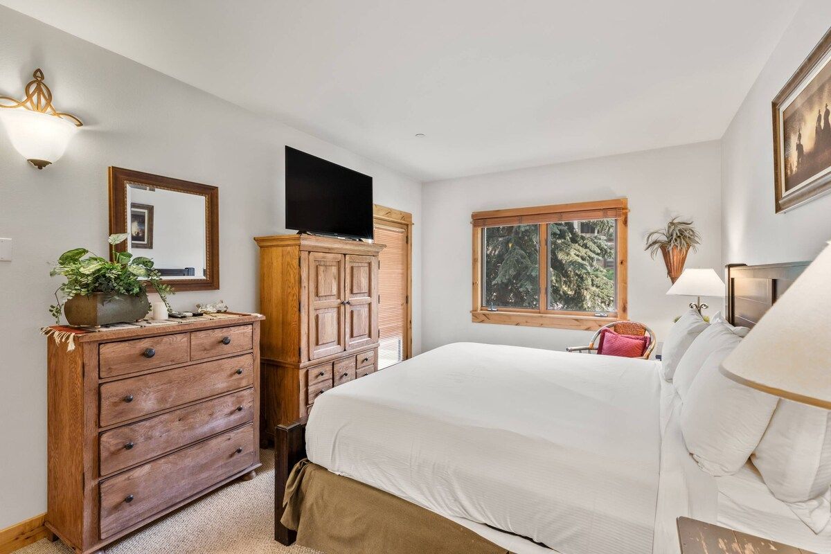 A bedroom with a bed, dresser, television, window, and a plant.