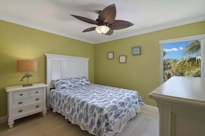 Ein Schlafzimmer mit Bett, Nachttisch, Kommode, Deckenventilator und einem Fenster mit Blick auf Palmen.