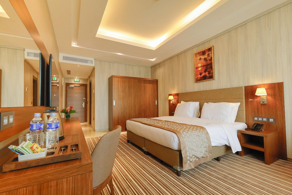 A hotel room with a large bed, a desk with a chair, a mirror, a nightstand with a lamp, and a wall-mounted artwork.