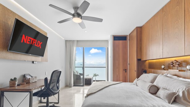 A modern hotel room with a bed, a desk, a chair, a TV, and a ceiling fan. - Zantamar 307B: Beachfront Suites w Ocean Views (Cruz de Huanacaxtle)