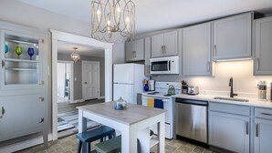 A modern kitchen with white cabinets, a stainless steel dishwasher, a microwave, and a gas stove. There is a dining area with a table and two stools.