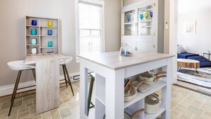 A modern kitchen with white cabinetry, a central island with pots, a dining area with a white table and bar stools, and a living area with a blue sofa and striped cushions.