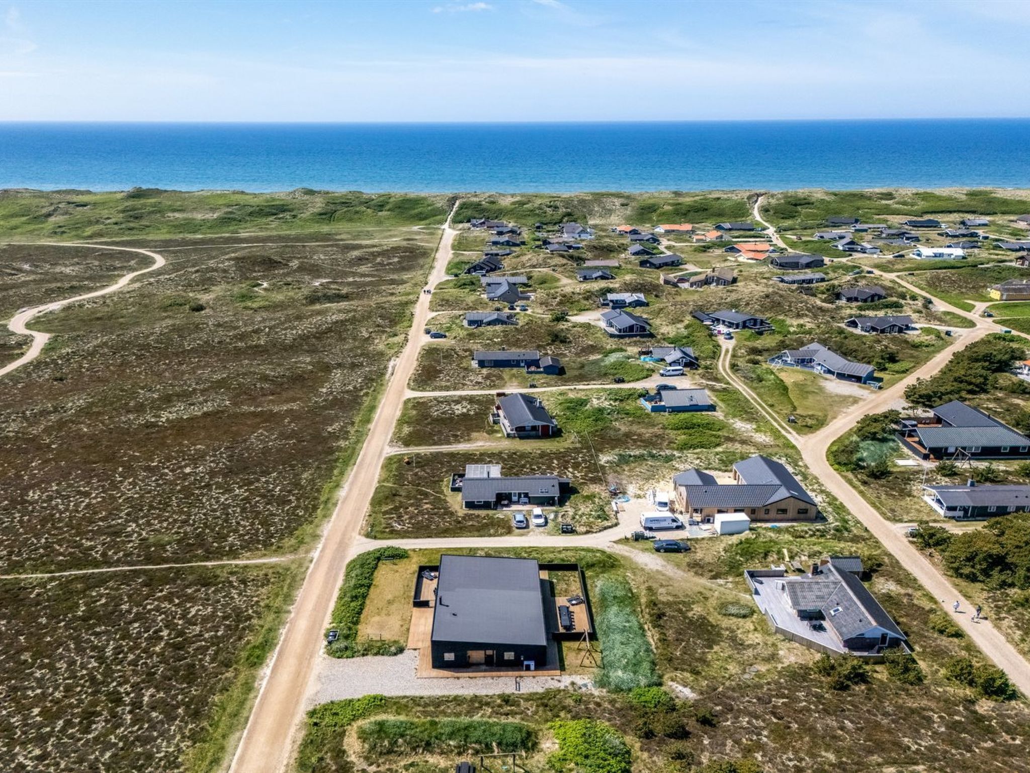 Vue aérienne d’une zone résidentielle côtière, avec des maisons, un bâtiment principal doté d’un toit sombre et une route menant à la mer.