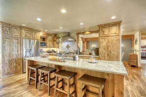 A spacious kitchen with wooden cabinets, a large island with stools, stainless steel appliances, and a stone backsplash.