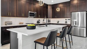 A modern kitchen with dark wood cabinets, stainless steel appliances, a central island with bar stools, and pendant lighting. - Vor4632td: 7 Bed, Bricktopia, Petting Zoo, Gaming Nook (Orlando)