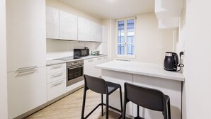 Una cocina moderna con armarios blancos, microondas, horno y cafetera. Hay dos taburetes negros y una ventana con persianas.