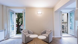 Un apartamento moderno con dos sillones grises y una pequeña mesa blanca, una lámpara en la pared y vistas a través de una puerta abierta.