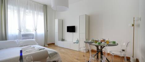 Ein Hotelzimmer mit einem Bett, einem Esstisch mit Stühlen, einem Fernseher und einem Fenster mit Vorhängen.