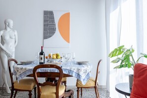 A dining area with a table set for four, a wine bottle, fruit, and a pitcher. A white statue is on the left, and a plant is on the right.