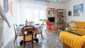 A living room with a yellow sofa, a wooden bookshelf, a dining table with chairs, and a TV. - GuestHost - Porta a Lucca - Pisa Bright Apartment with Balcony (Pisa)
