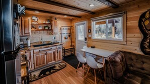 A cabin interior with wooden walls, a kitchen area, a dining table with chairs, and a large window. - Cozy Big Lake Cabin + Private Spa House, Hot Tub & Sauna (Wasilla)