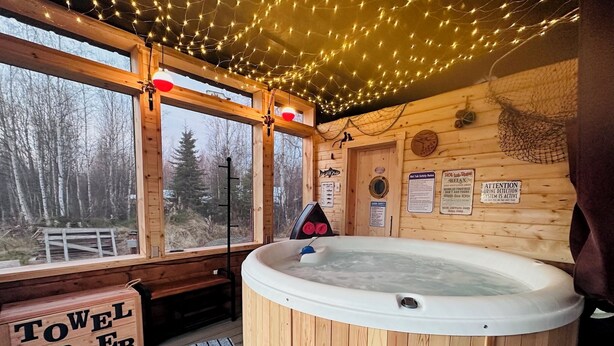 A wooden cabin with a hot tub, a towel warmer, and a view of the forest through large windows. - Cozy Big Lake Cabin + Private Spa House, Hot Tub & Sauna (Wasilla)