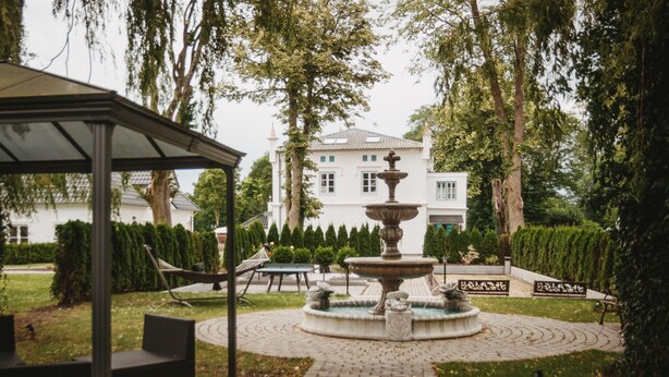 A gazebo, a fountain, and a white building with a garden in the background. - Kavaliershaus Erdgeschoss mit Eigener Terrasse im Henriettenhof (Grömitz)