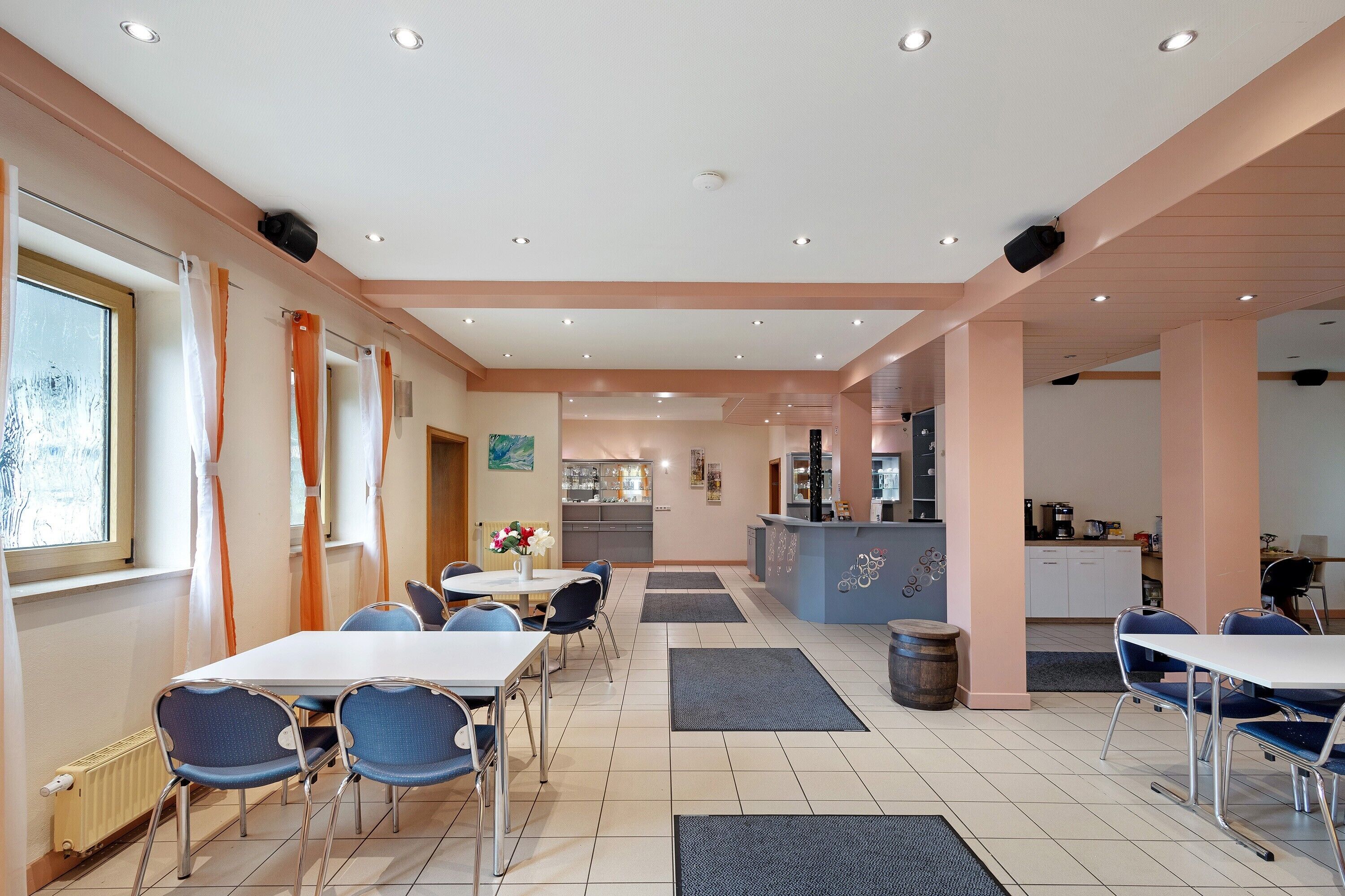 A hotel dining area with tables, chairs, a kitchen in the background, and a reception desk.
