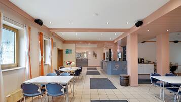 A hotel dining area with tables, chairs, a kitchen in the background, and a reception desk.