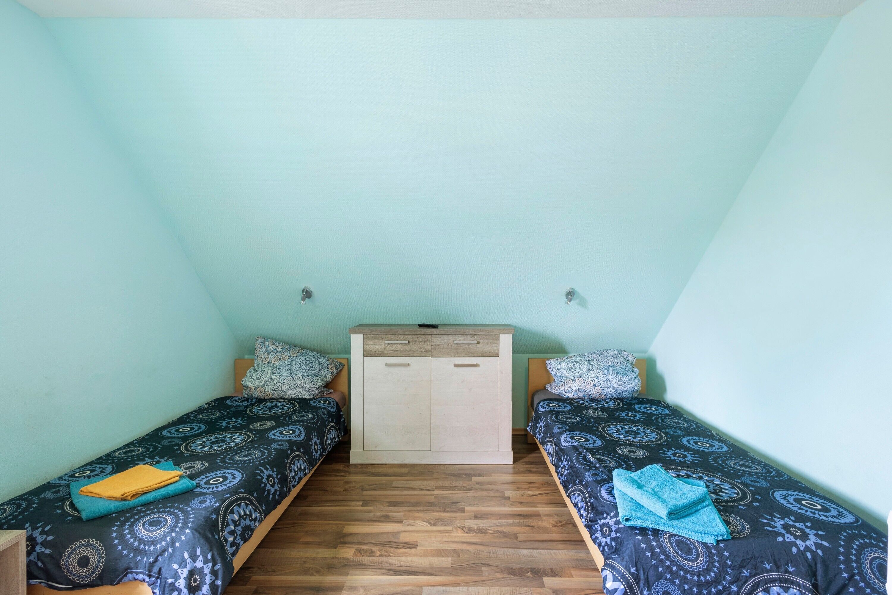 Une petite chambre à coucher composée d’une seule pièce, avec deux lits jumeaux, une armoire en bois et un mur de couleur claire.