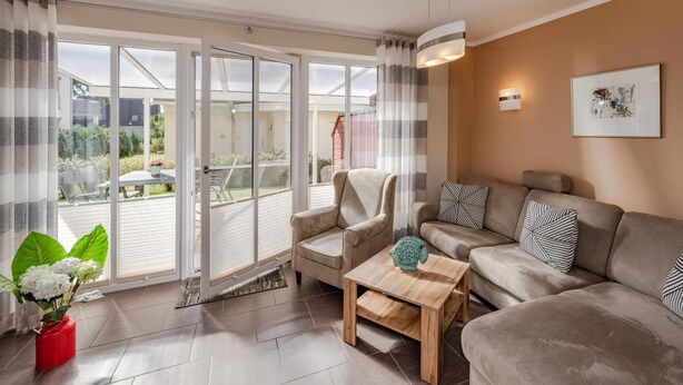 Ein Wohnzimmer mit einer Couch, einem Sessel und einem Couchtisch, einer Tür, die auf einen Balkon führt, und einem gerahmten Bild an der Wand.