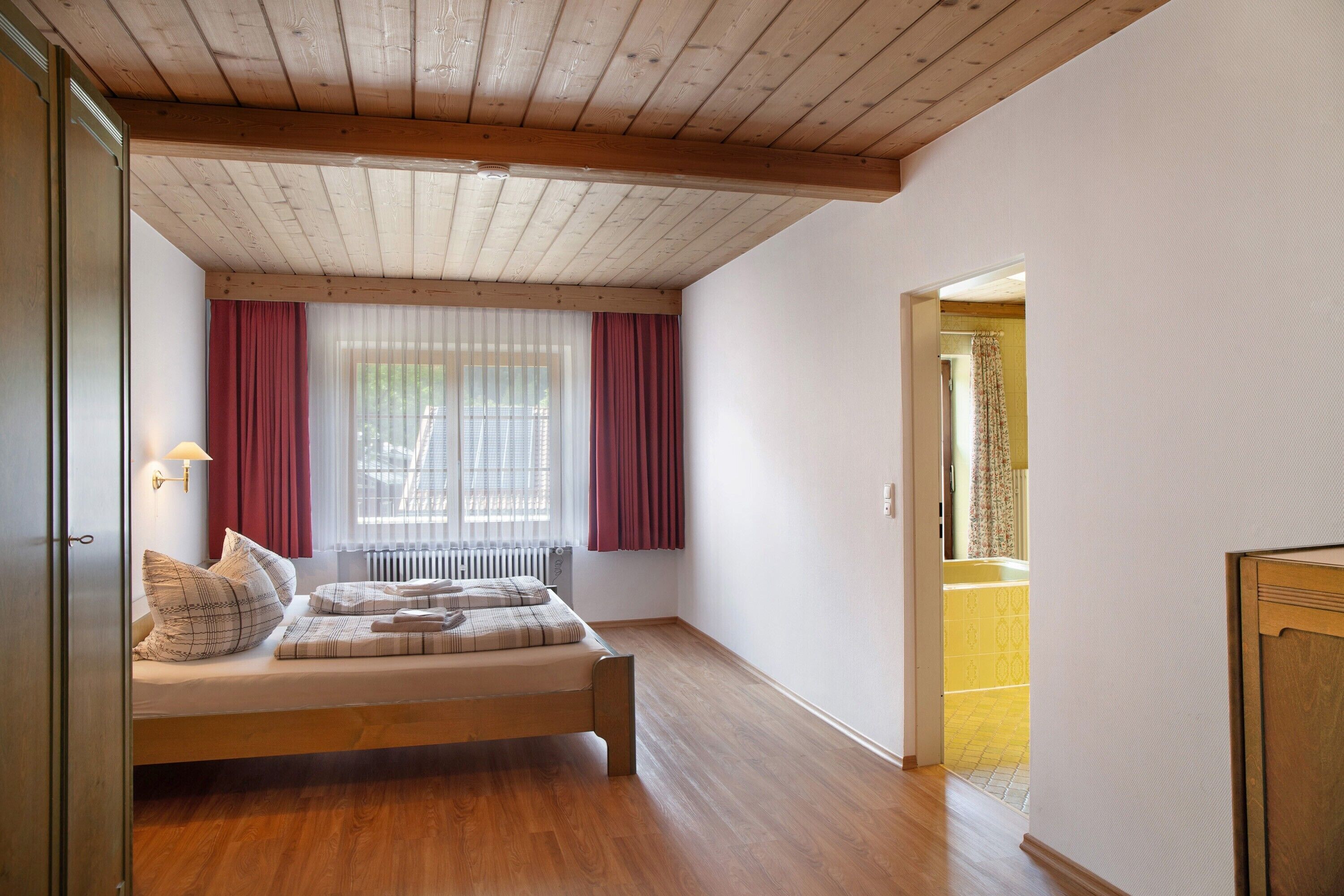 A bedroom with a wooden bed, a window with curtains, and a view into another room.