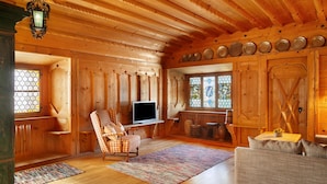 A wooden cabin interior with a television, a plaid armchair, a sofa, and a dining table.