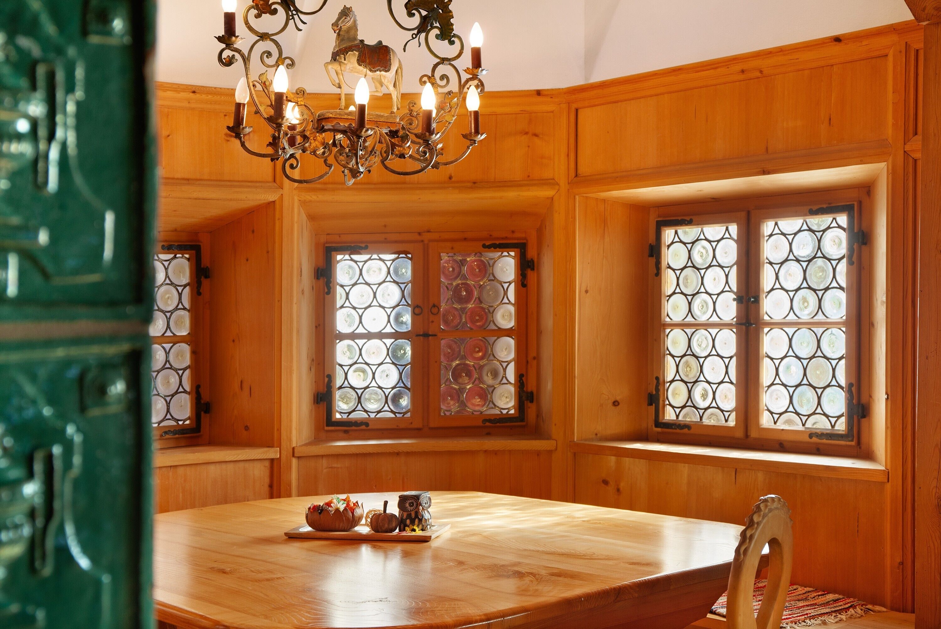 A wooden-paneled room with a chandelier, a table with a pumpkin decoration, and a window with a geometric pattern.