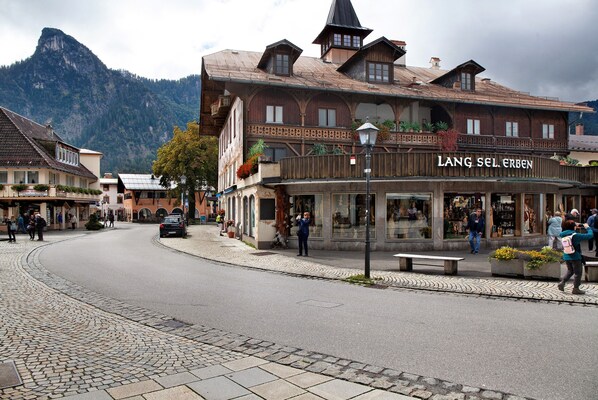 Ein traditionelles Holzhaus mit einem Schild, auf dem 'LANG SEL ERBEN' steht, auf einem Stadtplatz mit Kopfsteinpflaster und Bergen im Hintergrund.